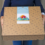 Eine Person hält eine große Schachtel von Grimm's mit dem Label Freies Spiel: Große Stufenpyramide, einem 100-teiligen Set bunter Holzbausteine, verziert mit gezeichneten Motiven.