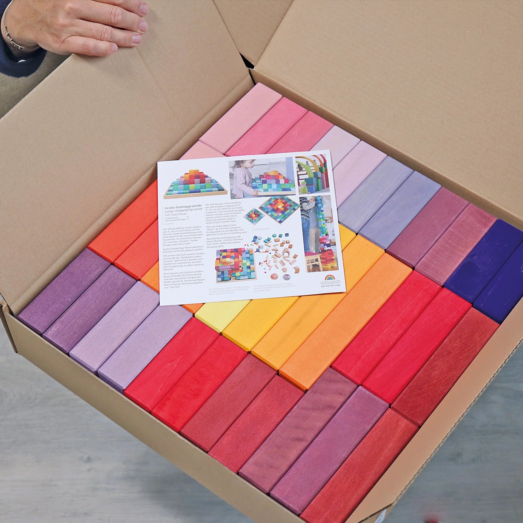 Freies Spiel: Große Stufenpyramide, eine Sammlung von rechteckigen Holzblöcken in Regenbogenfarben, liegt in einem Karton. Eine Anleitung zeigt kreative Stapelideen. Hände halten den Kartondeckel auf.
