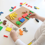 Hände eines Kindes beim Arrangieren von bunten Holzformen und Tierfiguren auf einer Korkplatte, Teil des Hämmerchenspiels, das handwerkliche Fähigkeiten fördert, hergestellt in einer geschützten Werkstätte in Deutschland.