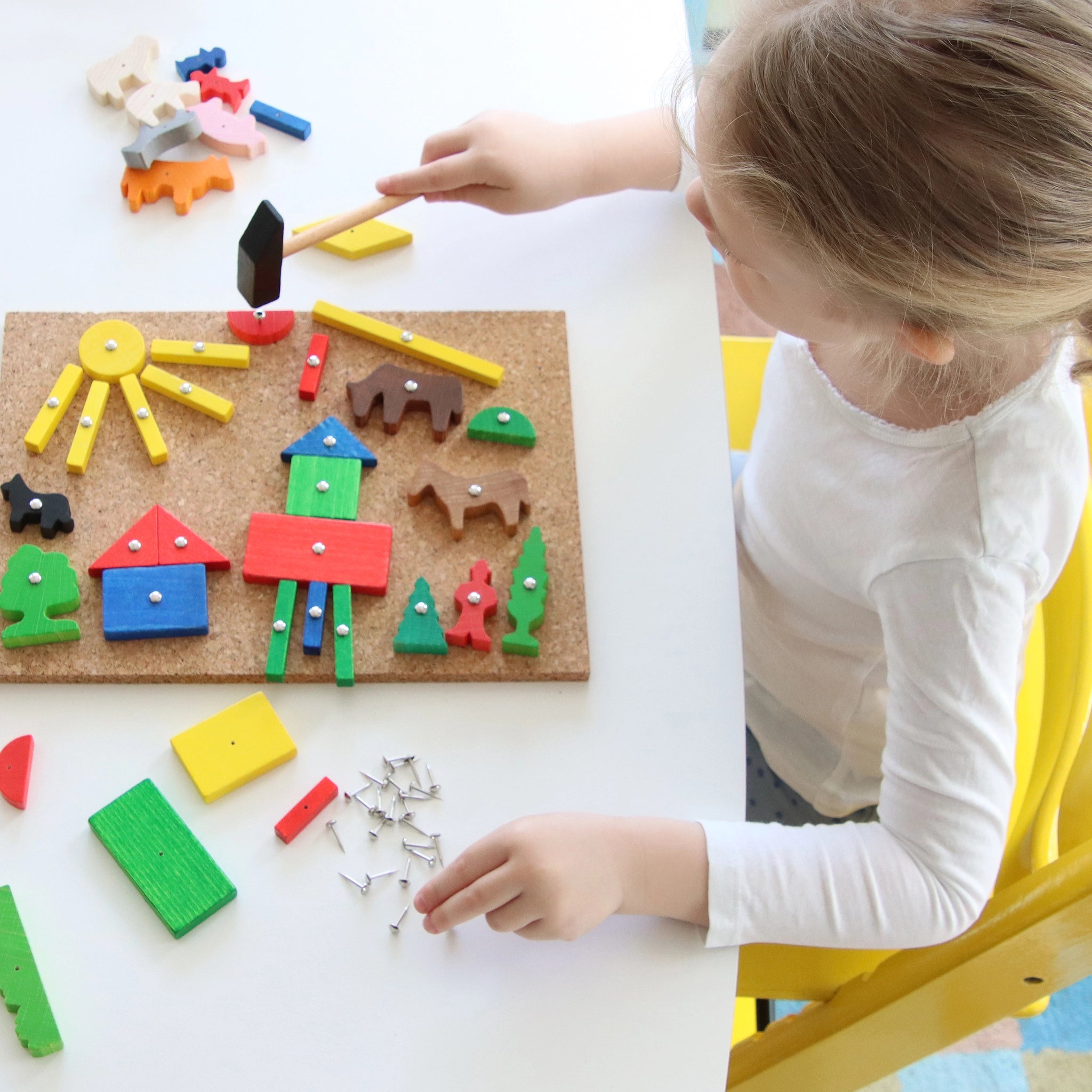 Ein Kind spielt am weißen Tisch mit dem Hämmerchenspiel, einem handgefertigten Set aus Kork und Holz. Es tackert bunte Formen mit einem Hämmerchen auf die Korkplatte, fördert Geschick und Kreativität.