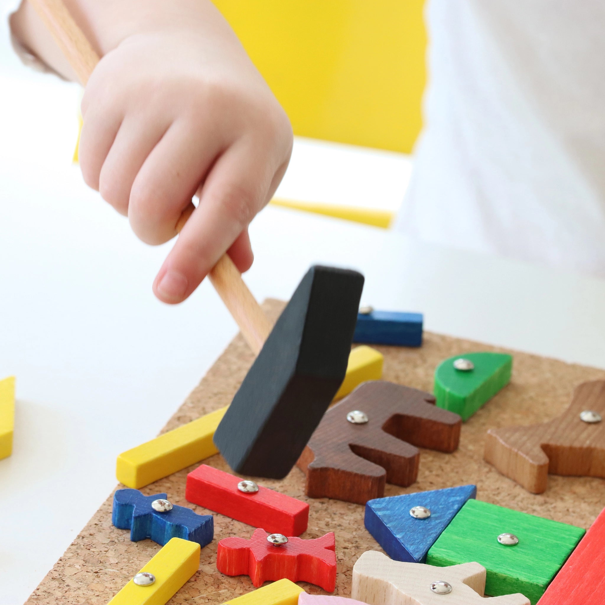 Ein Kind hält beim Hämmerchenspiel einen Holzhammer und hämmert farbenfrohe Holzformen auf ein Korkbrett. Dieses in Deutschland gefertigte Spielzeug unterstützt feinmotorische Fähigkeiten und handwerkliches Geschick.