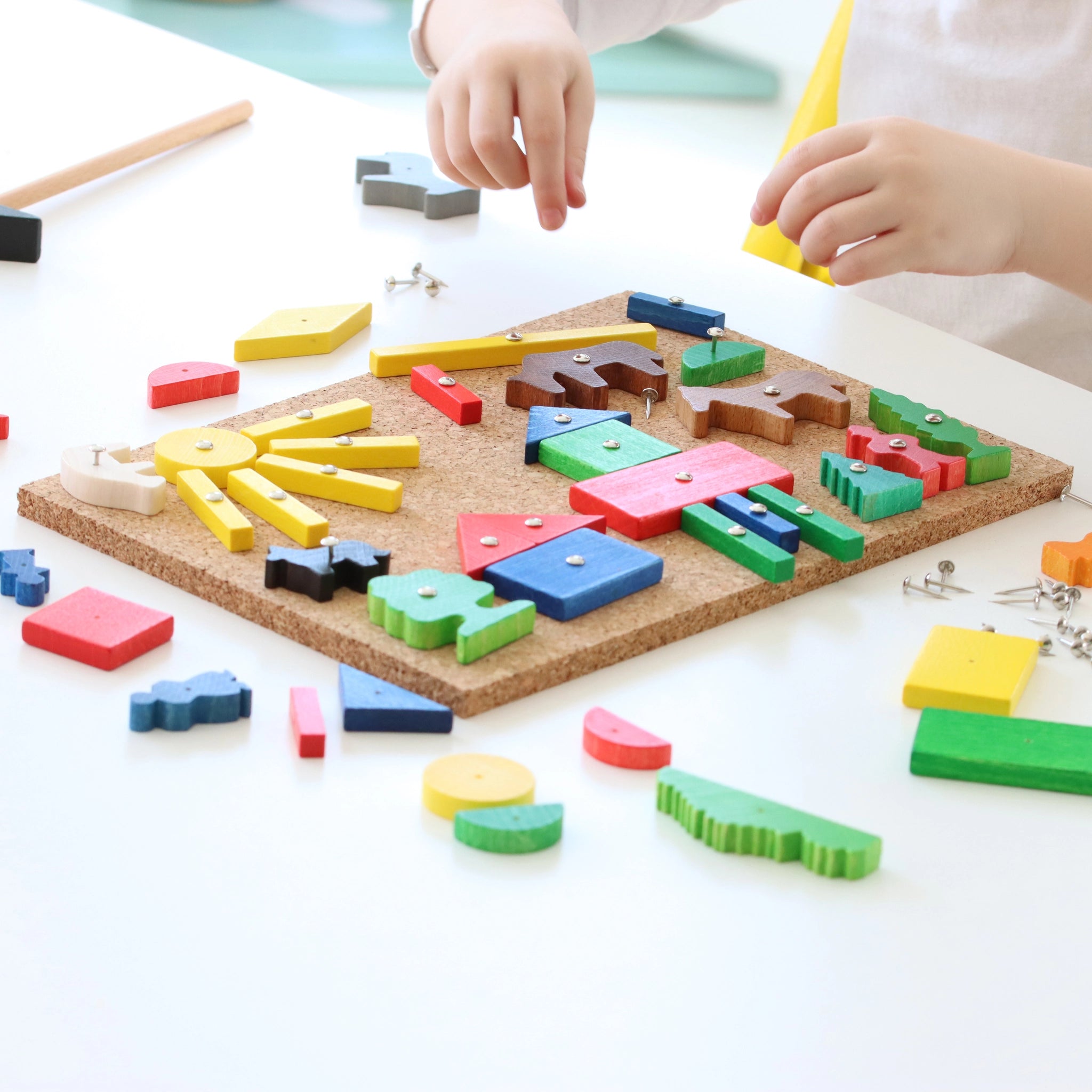 Hämmerchenspiel aus Kork und Holz: Ein Kinder-Bastelset auf einem Tisch, bestehend aus einem Korkbrett mit bunten Holzformen, die mit Nägeln fixiert werden. Kleine Hände positionieren Teile, ein Holzhammer liegt bereit.