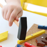Ein Kind hält einen kleinen Holzhammer über einem Korkbrett, auf dem das Hämmerchenspiel mit bunten Holzformen befestigt ist, ideal zur Förderung der Feinmotorik und handwerklichen Fähigkeiten.