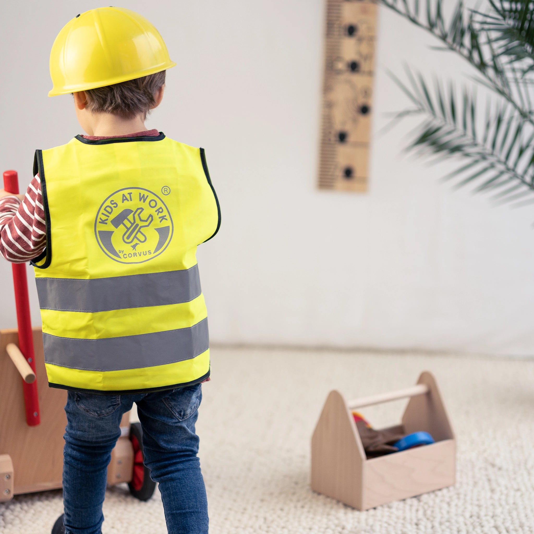 Kind trägt gelbe Weste und Bauhelm, hält Schubkarre aus massivem Buchenholz mit gummierten Rädern, ideal für kleine Handwerker. Produkt der Eigenmarke waelderspielzeug, gefertigt in Vorarlberg.