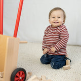 Schubkarre aus massivem Buchenholz, präsentiert von einem lächelnden Baby auf dem Boden. Räder mit hochwertigem Gummimantel, ideal für kleine Handwerker. Hergestellt in Vorarlberg, Österreich.