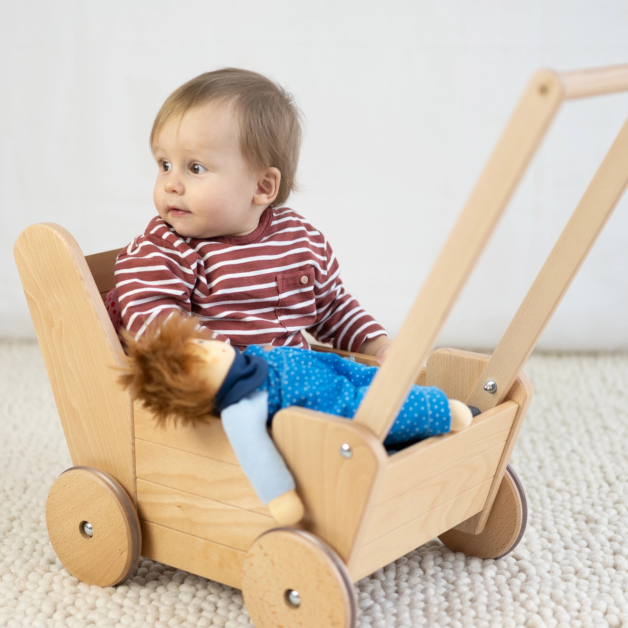 Ein Baby sitzt in einem stilvollen Puppenwagen aus massivem Buchenholz mit hochwertigen Gummirädern und stabiler Stahlachse, ausgestattet mit dreiteiligem Bettzeug, hergestellt in Vorarlberg.