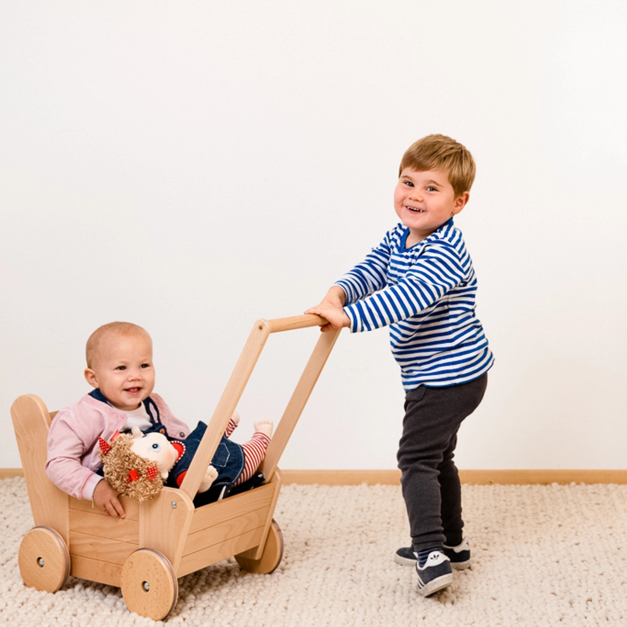 Junge schiebt Baby in einem Puppenwagen aus massivem Buchenholz mit hochwertigen Gummirädern und stabiler Stahlachse, inklusive 3-teiligem Bettzeug, hergestellt in Vorarlberg, Österreich.