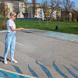 Frau in einem Parkplatz spielt Geschicklichkeitsspiel: Kugelfangspiel, aus massivem Buchenholz, fördert Geschicklichkeit und Koordination, hergestellt in Vorarlberg, Österreich, von waelderspielzeug.