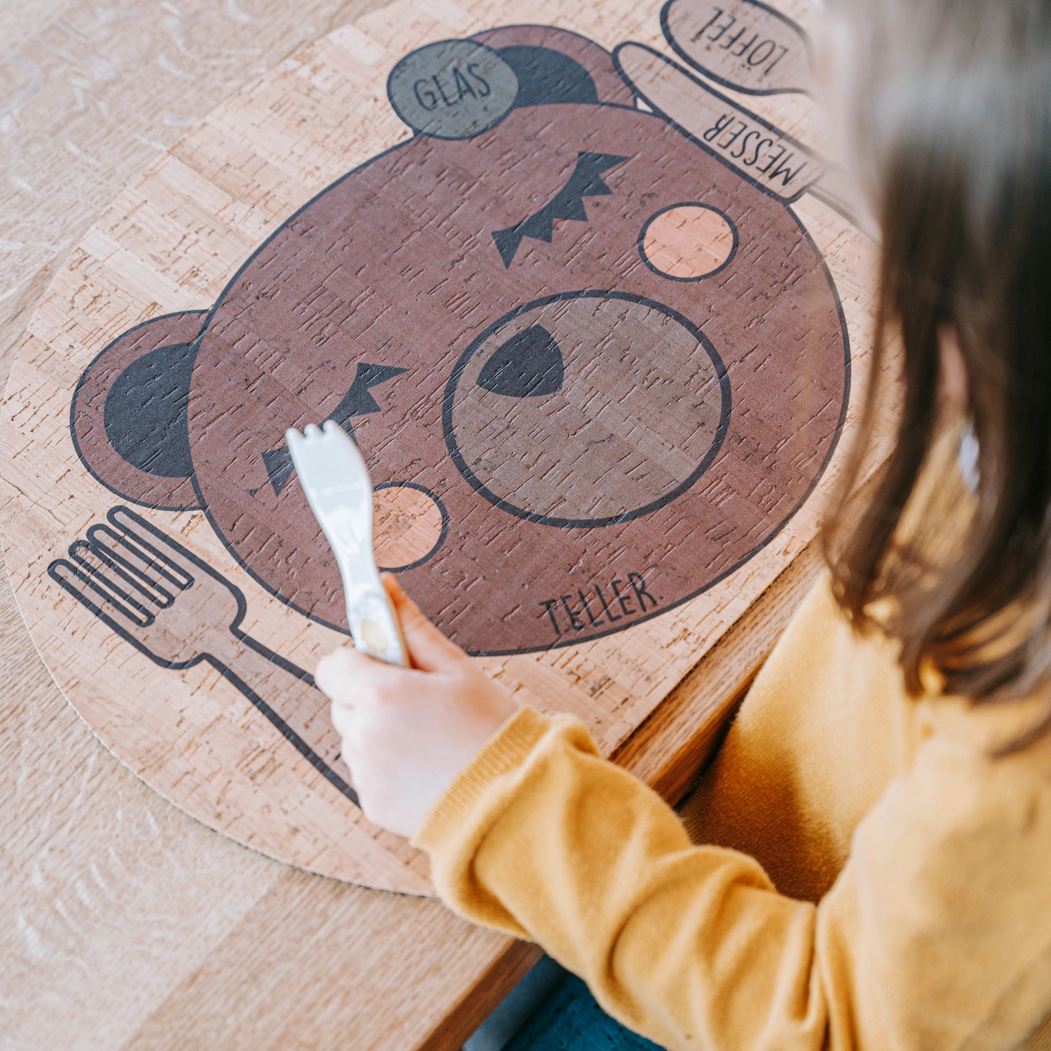 Kinder Tischset: Enjoying Bear aus Korkleder, inspiriert von Montessori, gehalten von einem Mädchen am Tisch. Hergestellt in Österreich, ideal für den Familientisch und fördert nachhaltiges Design.