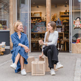 Zwei Frauen sitzen vor einem Laden auf kleinen Holzkisten, wobei der Kleiner Kinderhocker als stilvoller Beistelltisch dient. Hergestellt aus massivem Eschenholz, passend für zeitgemäße, nachhaltige Wohnräume.