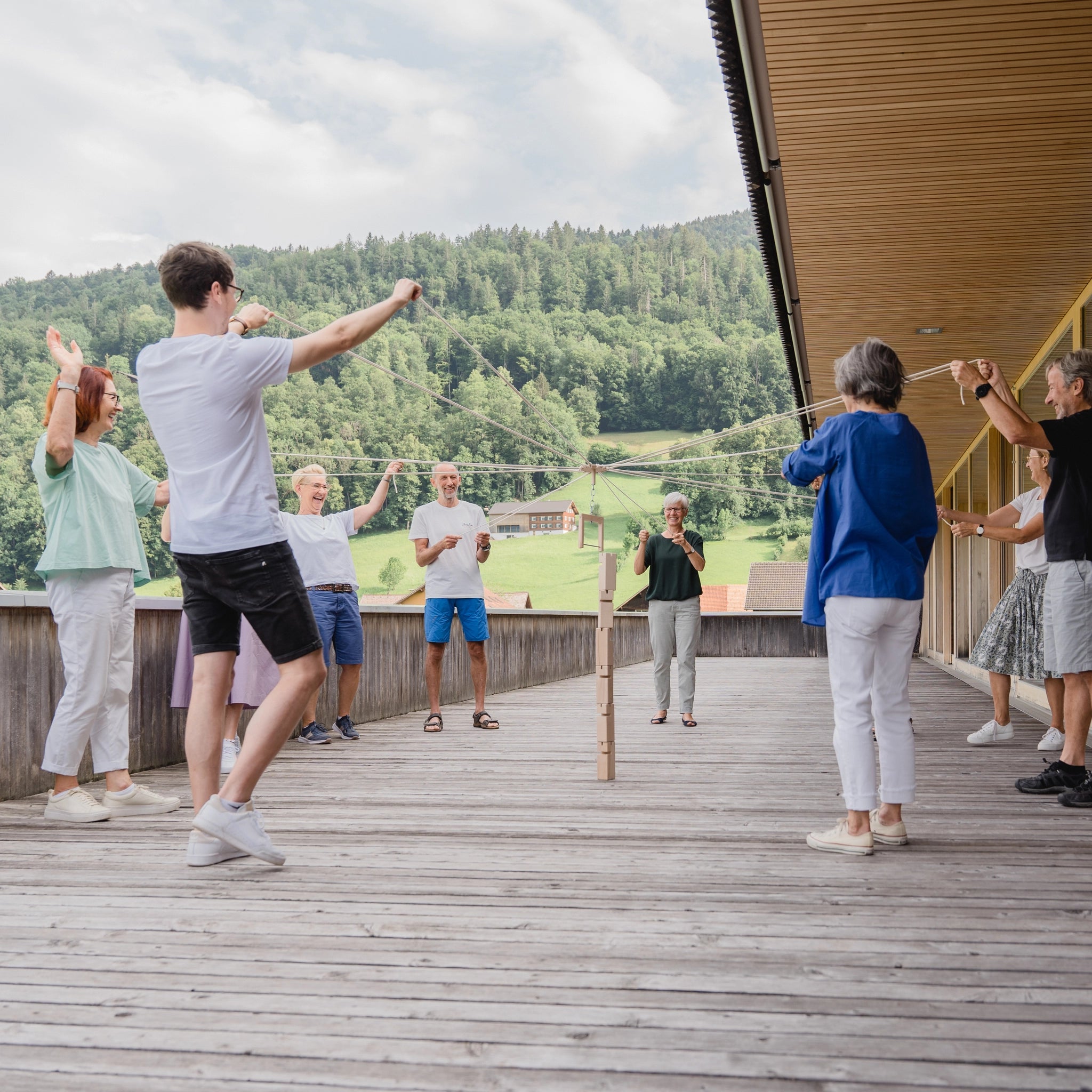 Teamspiel: Fröbelturm natur aus massivem Buchenholz, präsentiert auf einem Deck mit mehreren Personen im Hintergrund. Ideal für 4 bis 24 Spieler, erhältlich mit praktischem Transportbeutel, hergestellt in Vorarlberg.