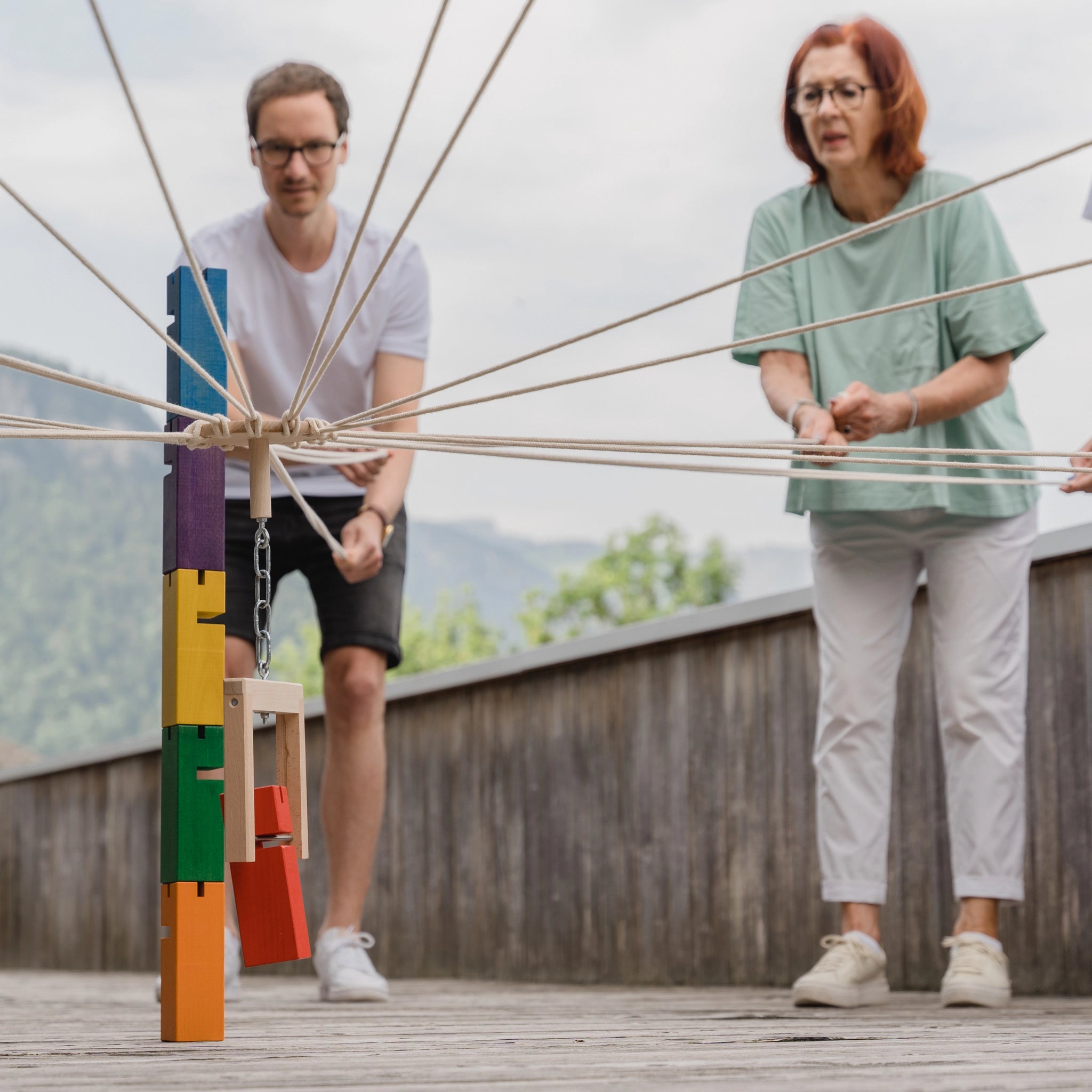 Teamspiel: Fröbelturm bunt aus massivem Buchenholz, dargestellt von Personen, die mit Seilen interagieren, geeignet für 4 bis 24 Spieler, erhältlich mit praktischem Transportbeutel, hergestellt in Vorarlberg, Österreich.