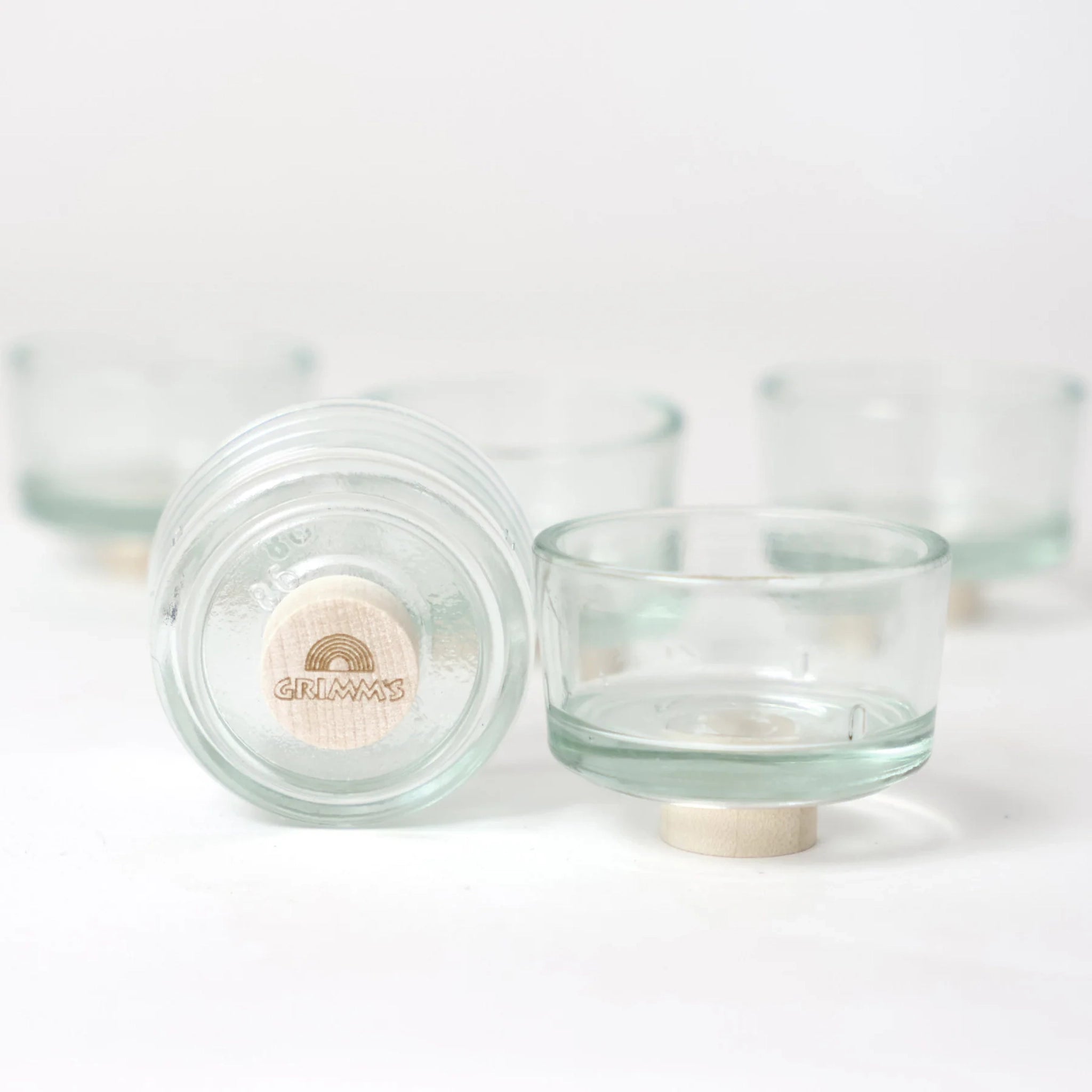 Steckfigur: Teelichthalter Glas, ein Set aus kleinen, klaren Glasschalen auf runden Buchenholzfüßen, eine zeigt das GRIMM'S Logo. Hergestellt in Deutschland, ideal für Snacks oder dekoratives Servieren.