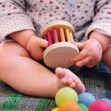 Kleinkind auf weicher Matte hält die Rassel: Regenbogen Mini-Babyroller aus Linden- und Buchenholz, umgeben von bunten Bällen. Fördert Greifen und Feinmotorik, nachhaltig in Deutschland produziert.