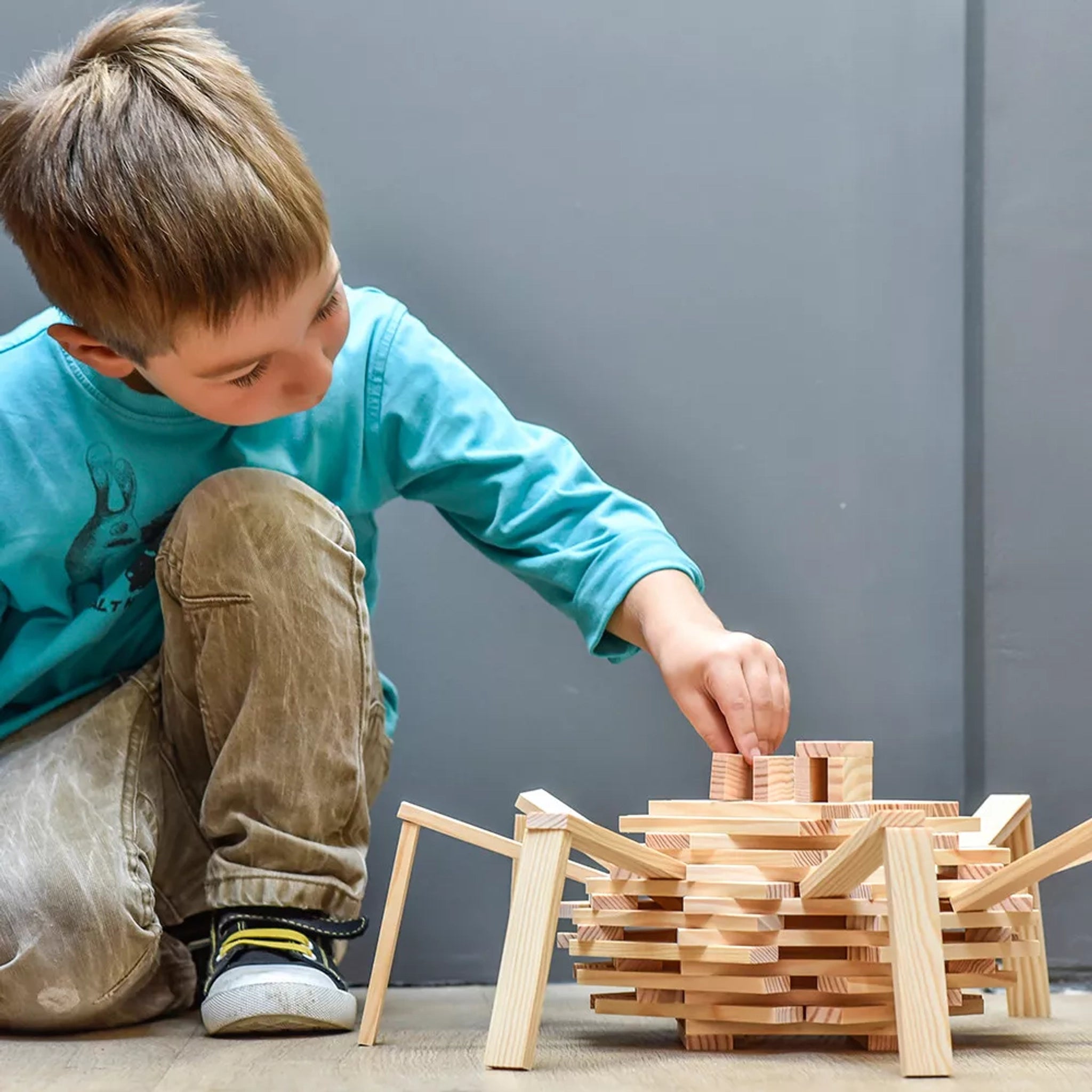 Junge beim Spielen mit Bausteine: 200er Box, baut Gitterstruktur aus naturfarbenen KAPLA-Plättchen, fördert Kreativität und räumliches Denken, hergestellt aus nachhaltigem Pinienholz in Frankreich.