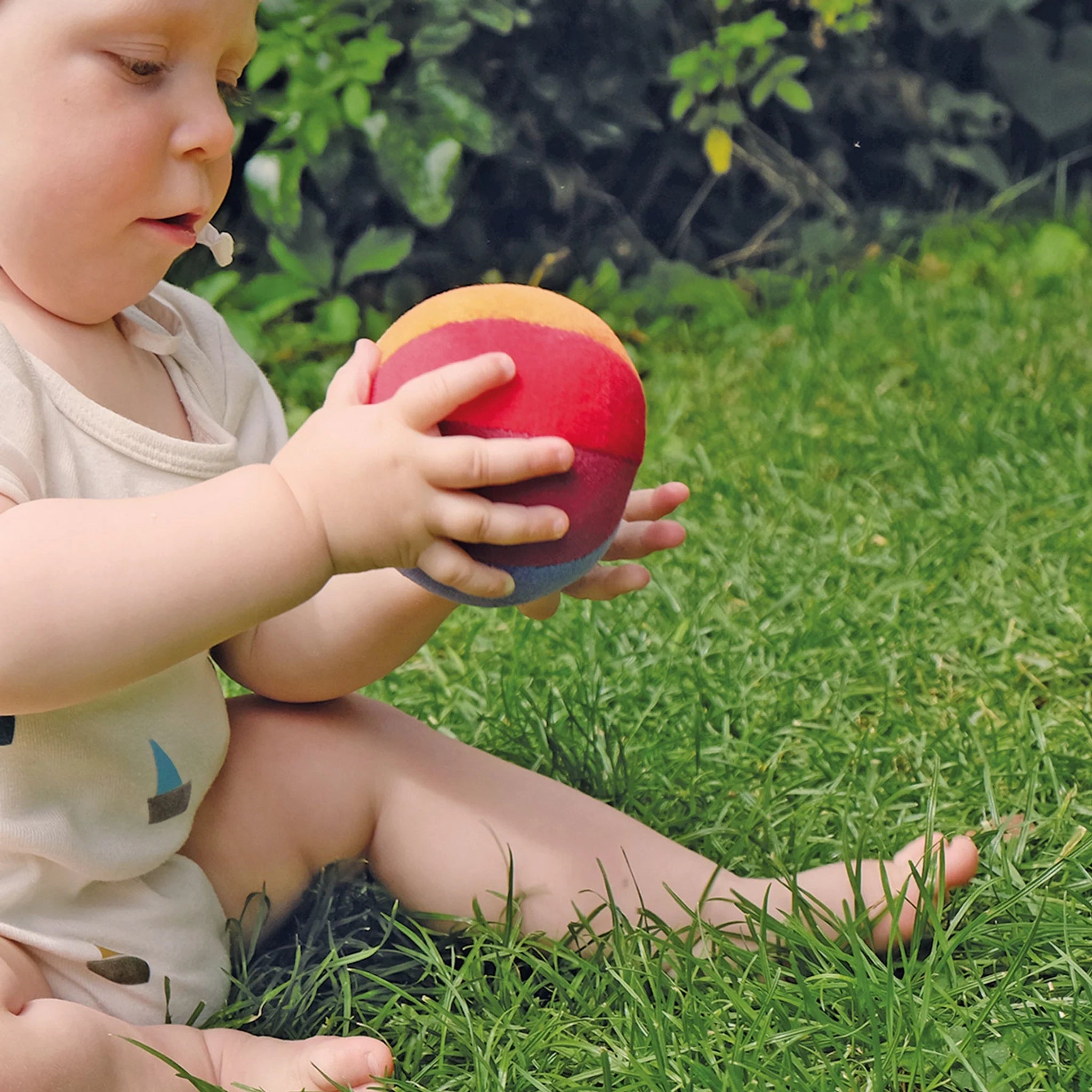 Baby sitzt auf Rasen, hält einen Regenbogenball, der sanft klingelt und Feinmotorik fördert. Der Ball, aus Nikistoff und Polyesterwolle, wird in einer deutschen Manufaktur nachhaltig gefertigt.