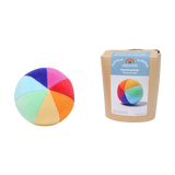 Der Regenbogenball aus weichem Nikistoff, bestehend aus acht bunten Segmenten, liegt neben seiner umweltfreundlichen Verpackung. Ein Glöckchen im Inneren fördert das Greifen und die Feinmotorik von Babys.