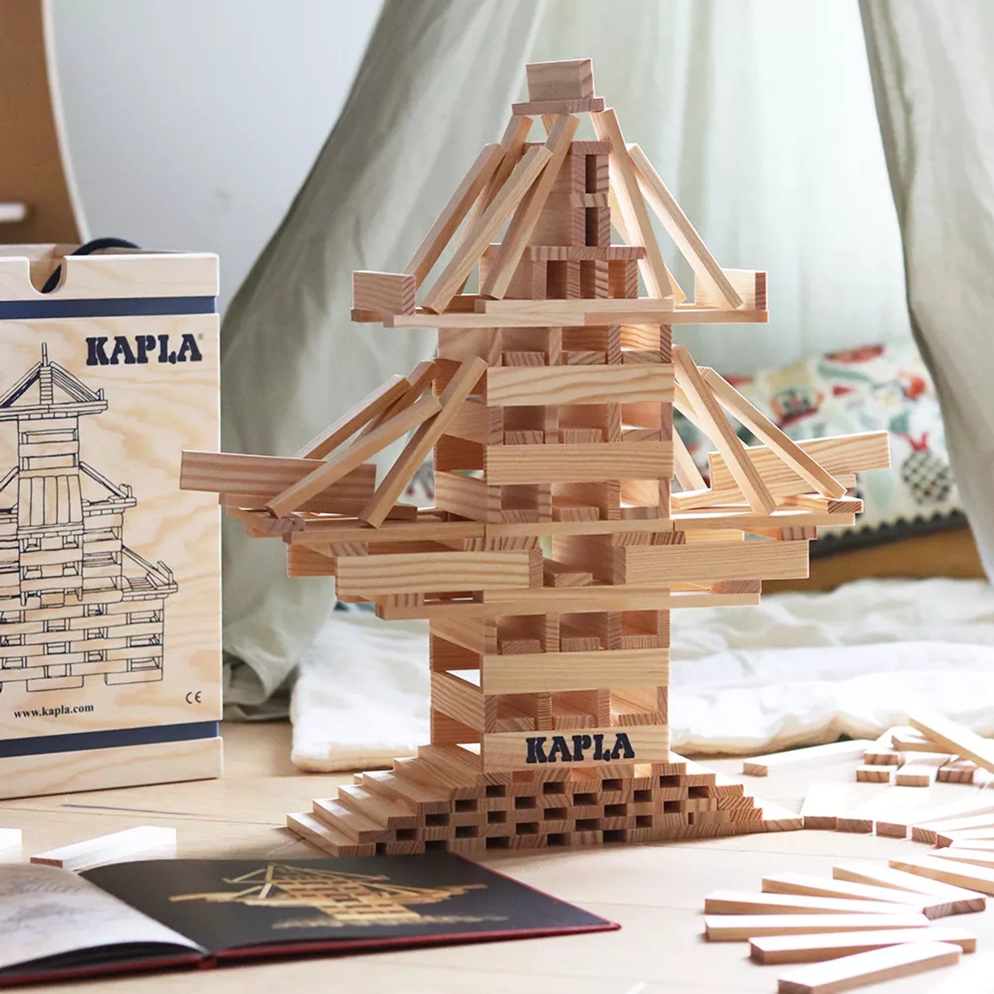 Bausteine: 280er Kasten zeigt einen Turm aus Kapla-Holzplättchen. Daneben sind eine Aufbewahrungsbox und ein Kunstbuch mit Bauideen sichtbar, um Kreativität und räumliches Denken zu fördern.