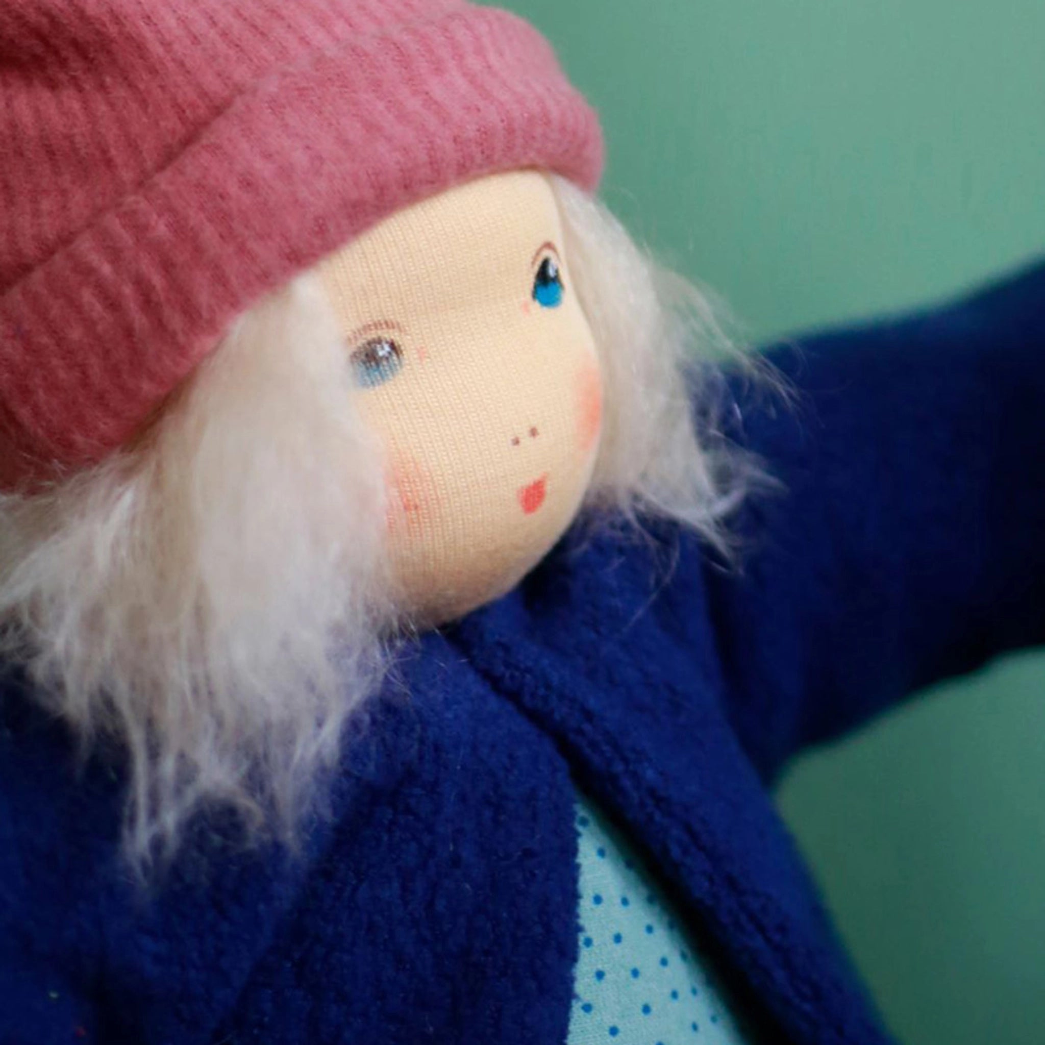 Anziehpuppe: Alma, eine handgemachte Stoffpuppe mit blonden Mohairhaaren, trägt eine blaue Jacke über einem hellblauen, gepunkteten Outfit. Gefertigt aus Bio-Baumwolle, mit schadstofffreier Schafschurwolle gefüllt.