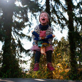 Ein junges Kind springt mit der Anziehpuppe: Alma, einer weichen Bio-Baumwollpuppe mit Mohairhaaren, über einem Trampolin. Alma trägt ein abnehmbares Kleid, Höschen und Mütze.