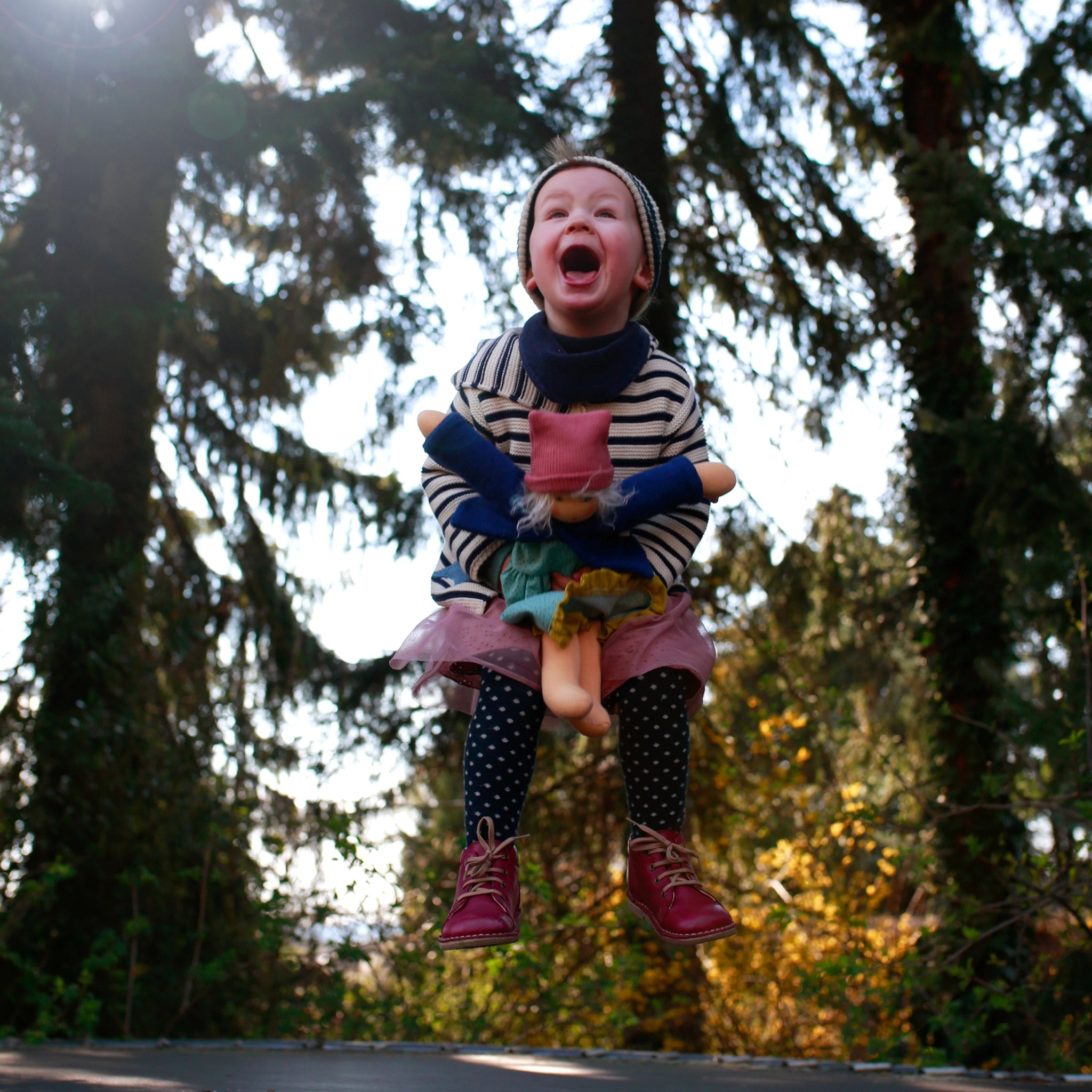 Ein junges Kind springt mit der Anziehpuppe: Alma, einer weichen Bio-Baumwollpuppe mit Mohairhaaren, über einem Trampolin. Alma trägt ein abnehmbares Kleid, Höschen und Mütze.