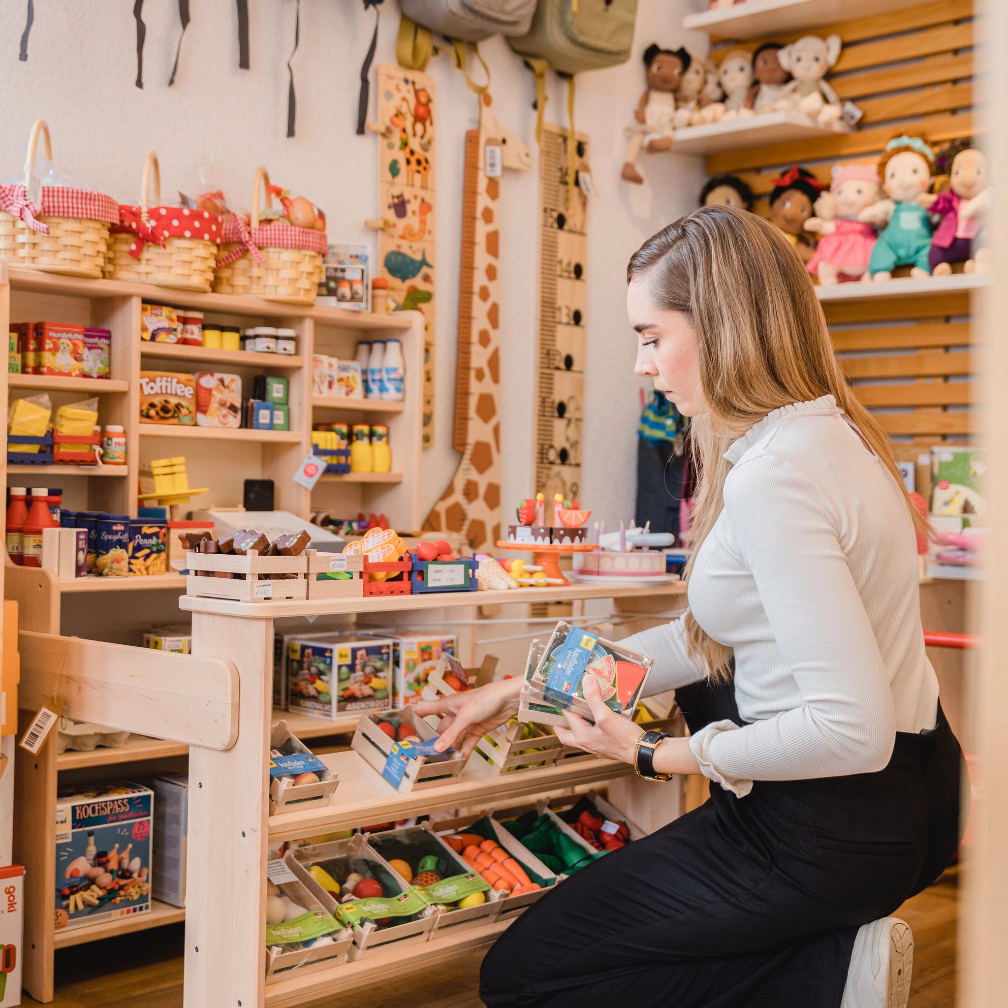 Frau im Spielzeugladen hält Lebensmittel: Süßwaren klein, ein Set aus 5 bunten Süßigkeiten aus robustem Buchenholz, präsentiert in einer kleinen Holzkiste, hergestellt in deutscher Holzmanufaktur.