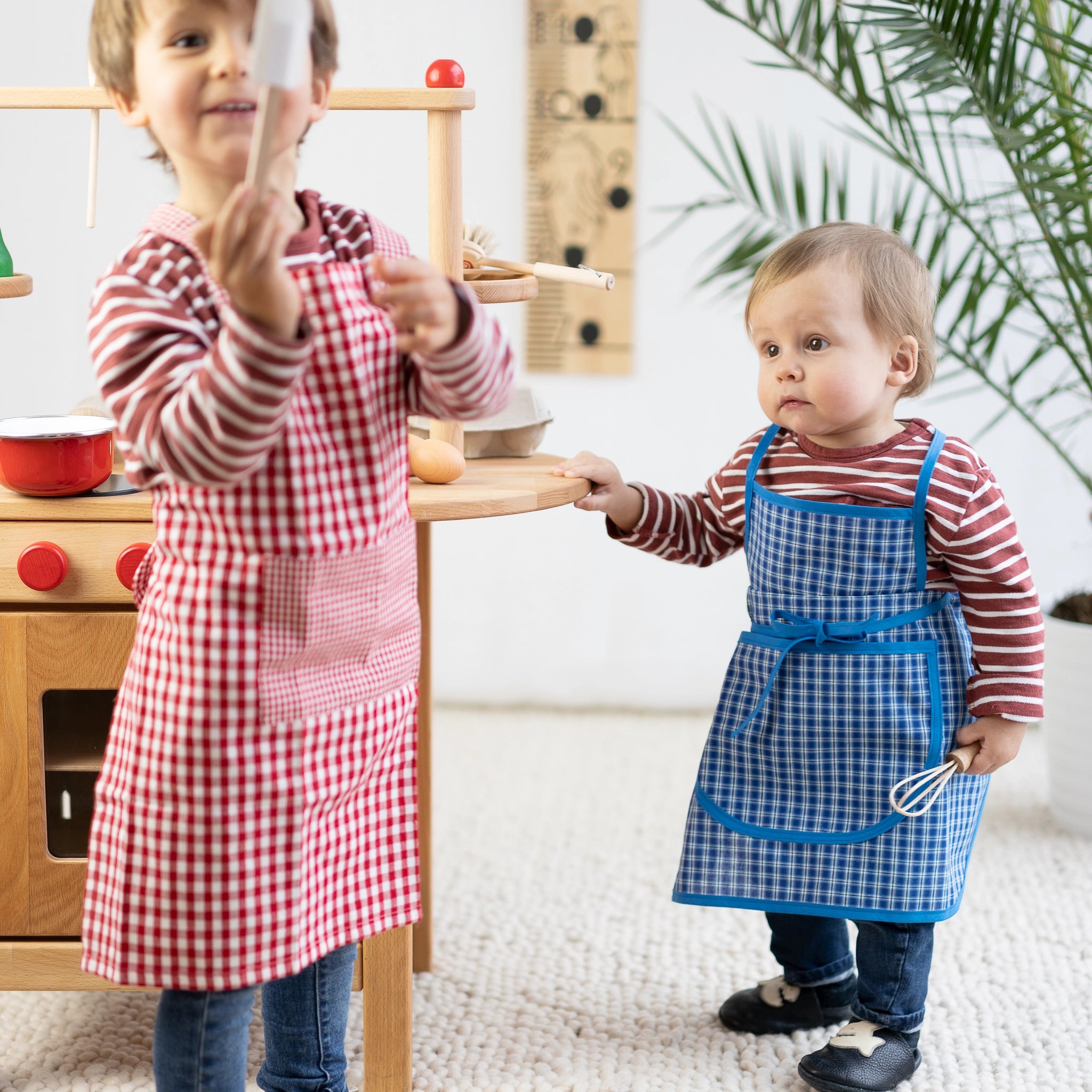 Zwei Kinder in einer Küche tragen Schürzen und verwenden den Kinderschneebesen. Dieser hochwertige Schneebesen aus unbehandeltem Buchenholz ist perfekt für kleine Köche, um mitzuhelfen.