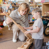 Frau und Junge spielen mit dem Holzfahrzeug: Mobilkran. Der robuste Kran aus Buchenholz hat einen drehbaren, höhenverstellbaren Kranarm und wird mit zwei Kranführerfiguren geliefert.