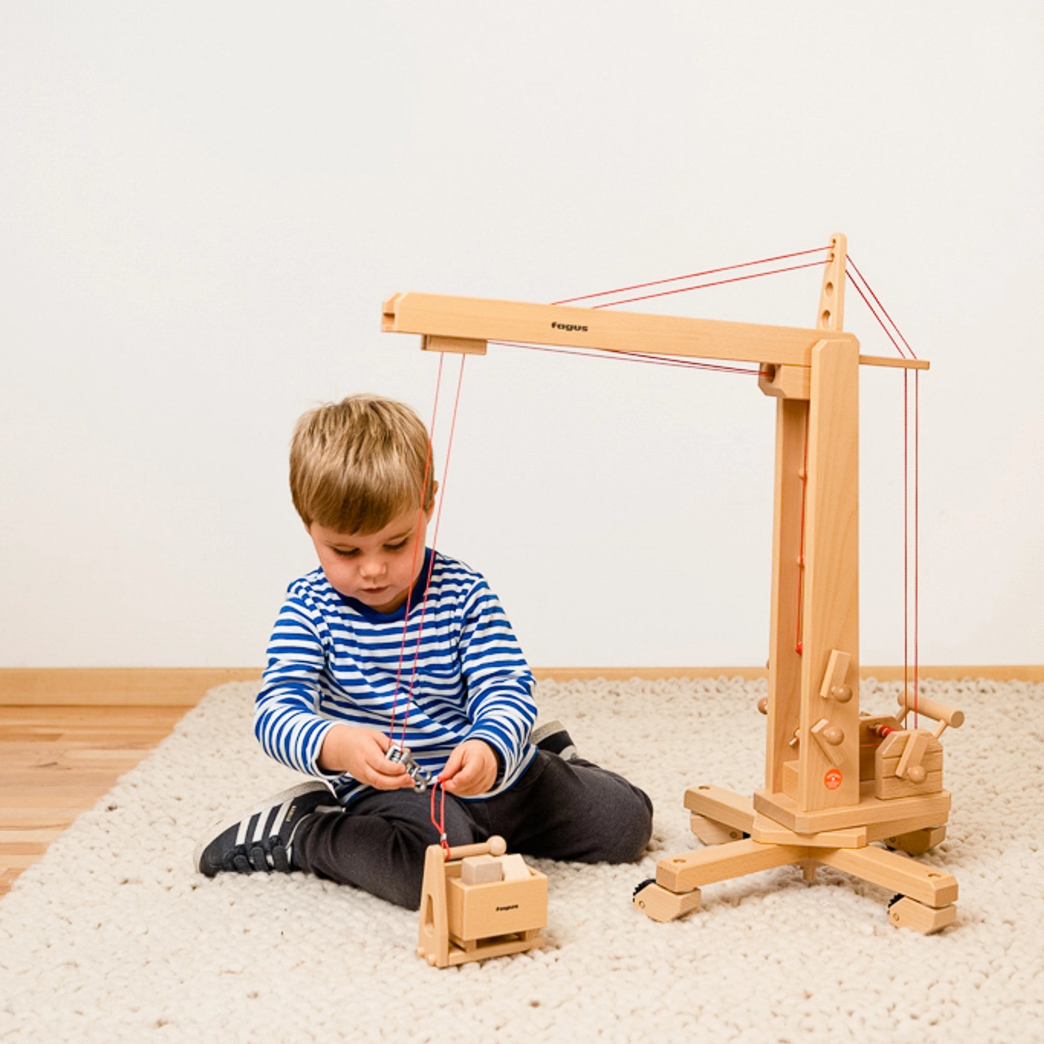 Ein Junge spielt auf dem Boden mit dem Holzfahrzeug: Hochkran aus massivem Buchenholz, dessen Turm drehbar ist und dessen Ausleger höhenverstellbar mittels Kurbeln ist. Ideal für kreatives, technisches Spiel.