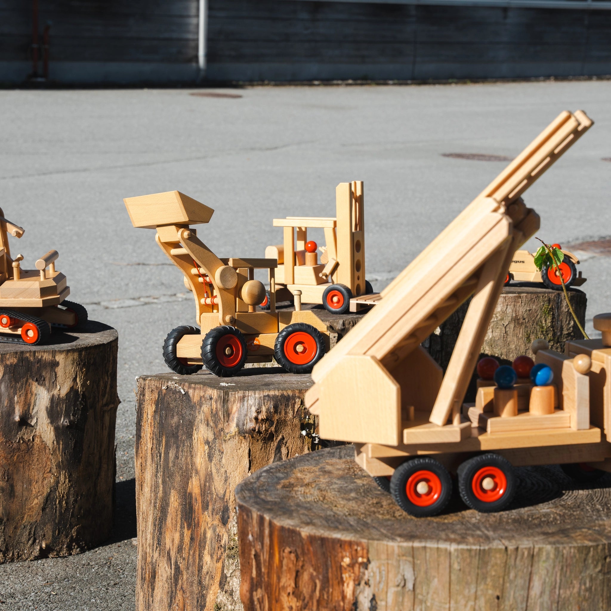 Holzfahrzeug: Radlader aus massivem Buchenholz, auf Baumstümpfen präsentiert. Mit verstellbarem Ausleger und Schaufel, inklusive zwei Fahrerfiguren. Handgefertigt in Deutschland, symbolisiert nachhaltige Produktion und soziales Engagement.