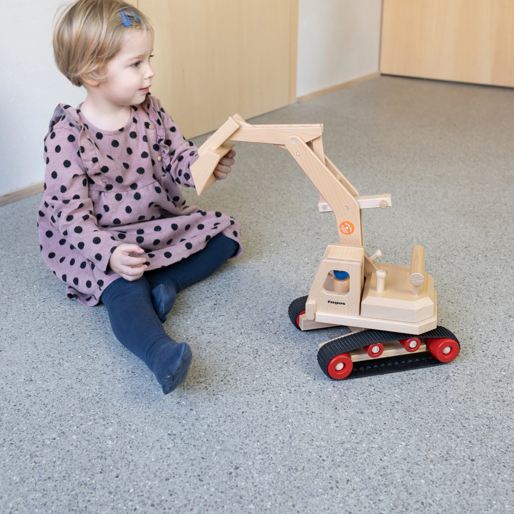 Ein Kind spielt mit dem Holzfahrzeug: Bagger aus massivem Buchenholz, mit drehbarem Aufbau und beweglichem Schaufelarm. Inklusive zwei Fahrerfiguren und stabilen Gummiketten, hergestellt in Deutschland.