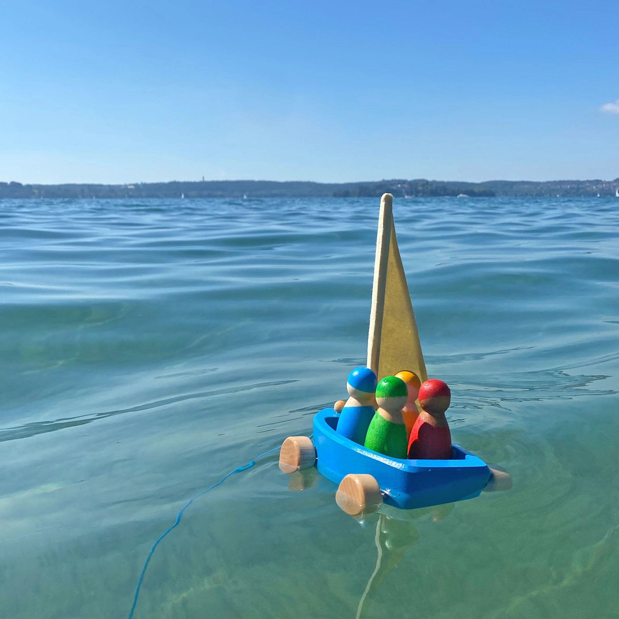 Holzfahrzeug: Strandsegler, ein schwimmfähiges Holzspielzeug-Set mit Segelschiff und vier Matrosen, im Wasser treibend. Ideal für drinnen und draußen, hergestellt aus lasiertem Lindenholz in Deutschland.