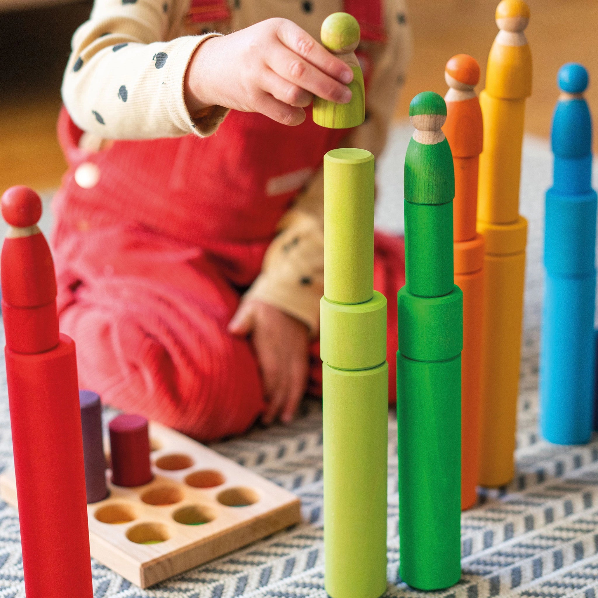 Freies Spiel: Regenbogenfreunde – ein Kind spielt drinnen mit bunten Holzfiguren. Das Set aus 12 handbemalten Figuren fördert kreatives Spielen und wird in Deutschland nachhaltig hergestellt.