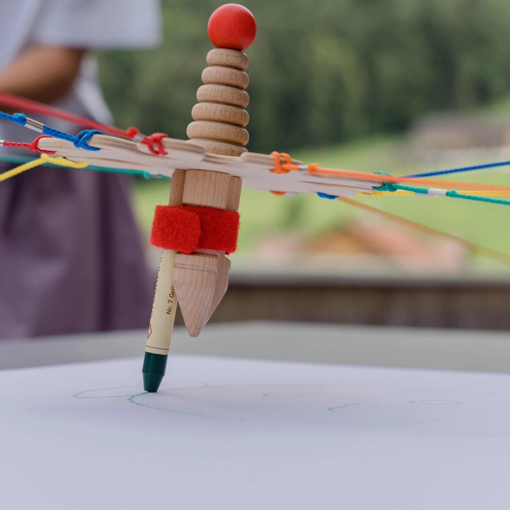 Teamspiel: Strippenzieher, ein Holzspielzeug mit roter Kugel oben, auf einem Blatt Papier präsentiert, geeignet für drinnen und draußen. Produziert in Zusammenarbeit mit der Karl-Schubert-Werkstätte.