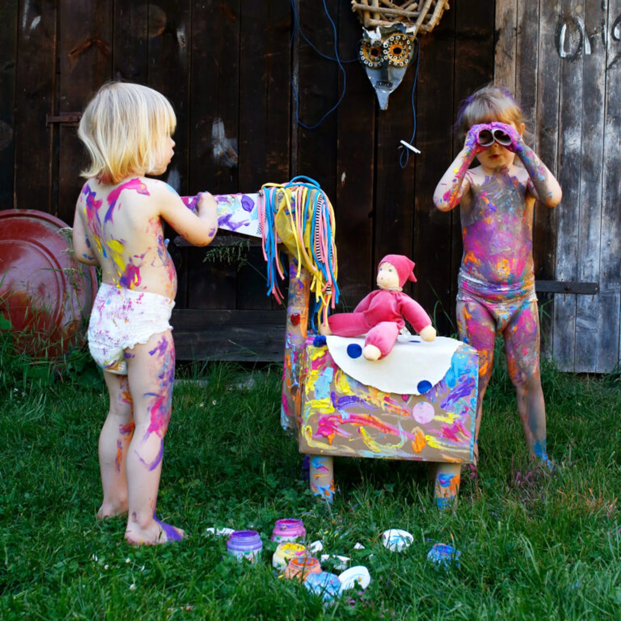 Zwei Kinder spielen mit Farbe, während die Puppe Nani pink mit süßem Bubi-Kragen und frecher Mütze aus weichem Bio-Baumwollnicki und schadstofffreier Schafschurwolle im Vordergrund sitzt.