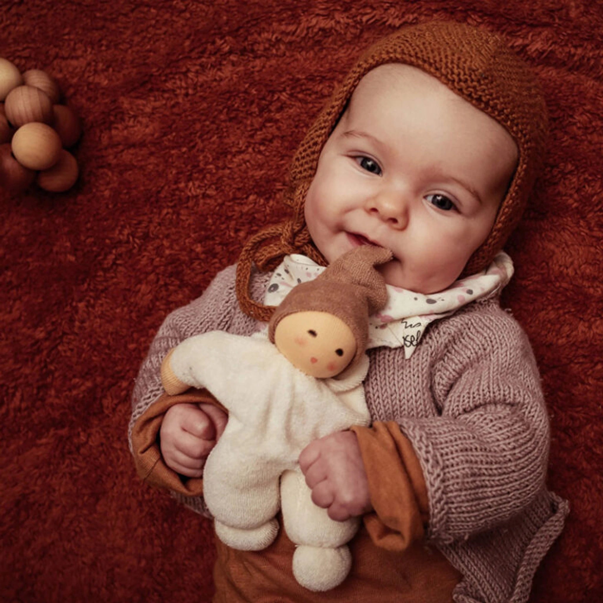 Ein Baby liegt auf einem roten Teppich und hält die Puppe: Nucki beige, gefertigt aus weichem Bio-Baumwollefrottee mit sanft klingelndem Glöckchen, ideal zum Greifen und Kuscheln.