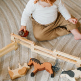 Holzzaun: Klappbar, aus massivem Kieferholz, ideal für das Spielen mit Holztieren. Ein Baby spielt mit den Holzspielzeugen auf einer Decke, was die Vielseitigkeit des Zauns unterstreicht.