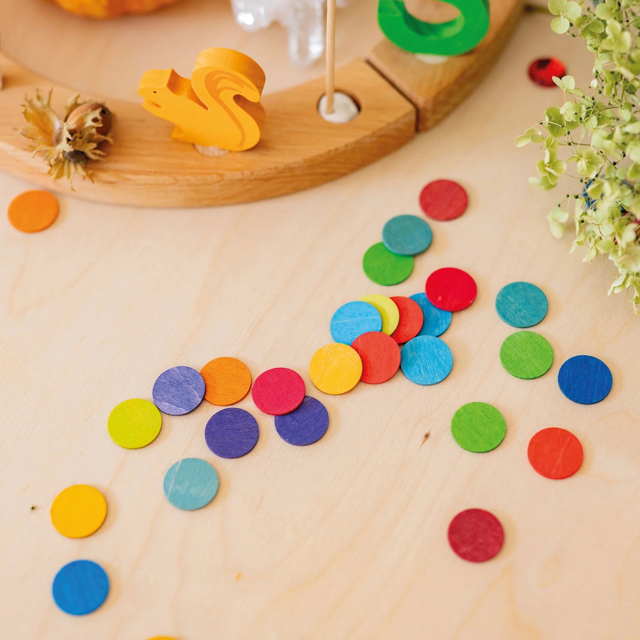 Freies Spiel: Konfettitaler Regenbogen, bunte Holzscheiben auf hellem Untergrund, kombiniert mit einem Festtagsring und einem orangen Eichhörnchen, ideal für festliche Tischdekorationen. Hergestellt in einer deutschen Manufaktur.