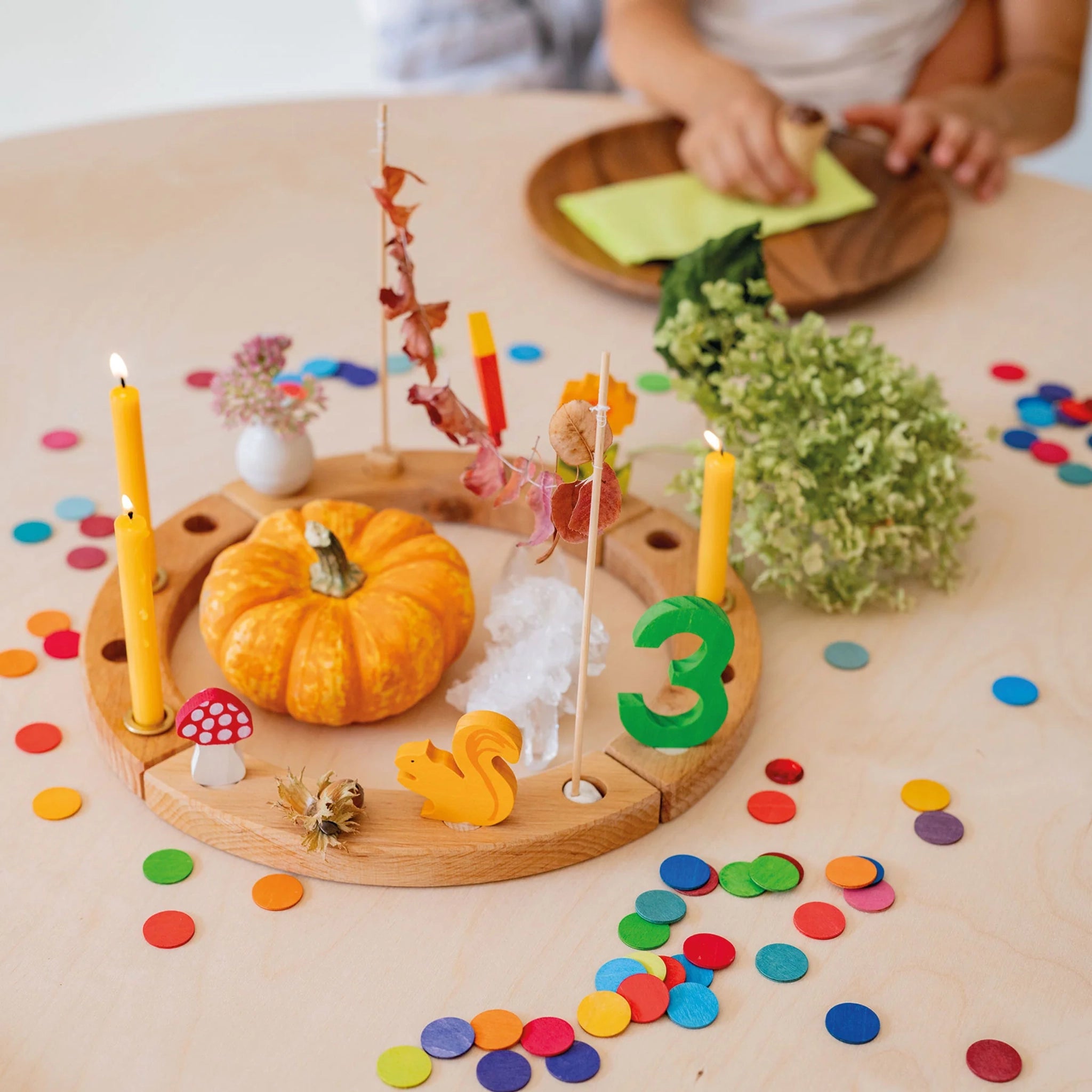 Freies Spiel: Konfettitaler Regenbogen dekorieren einen Tisch mit farbenfrohen Sperrholzkonfettitalern, umgeben von schlanken gelben Kerzen, einem Miniaturkürbis, Holzfiguren und getrockneten Blättern, ideal für festliche Anlässe.
