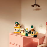 Bauwelt: Movement, ein abstrakter Baukasten aus farblich lasiertem Lindenholz, präsentiert sich auf einem pastellrosa Sideboard. Holzbausteine in vielfältigen Formen laden zum experimentellen Bauen ein.