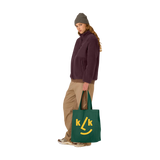 Eine Frau hält eine Stofftasche Tannengrün aus 100% recycelter Baumwolle mit gelbem KLICKKLACK-Design. Die Tasche hat Balgfalten für Volumen und lange Tragegriffe.