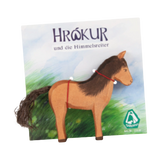 Holztier: Islandpferd Hrókur vor einem stilisierten Karton mit dem Titel Hrö́kur und die Himmelsreiter, verziert mit einer roten Zügel und einem Garnschwanz, von Ostheimer, Art.-Nr. 11115.