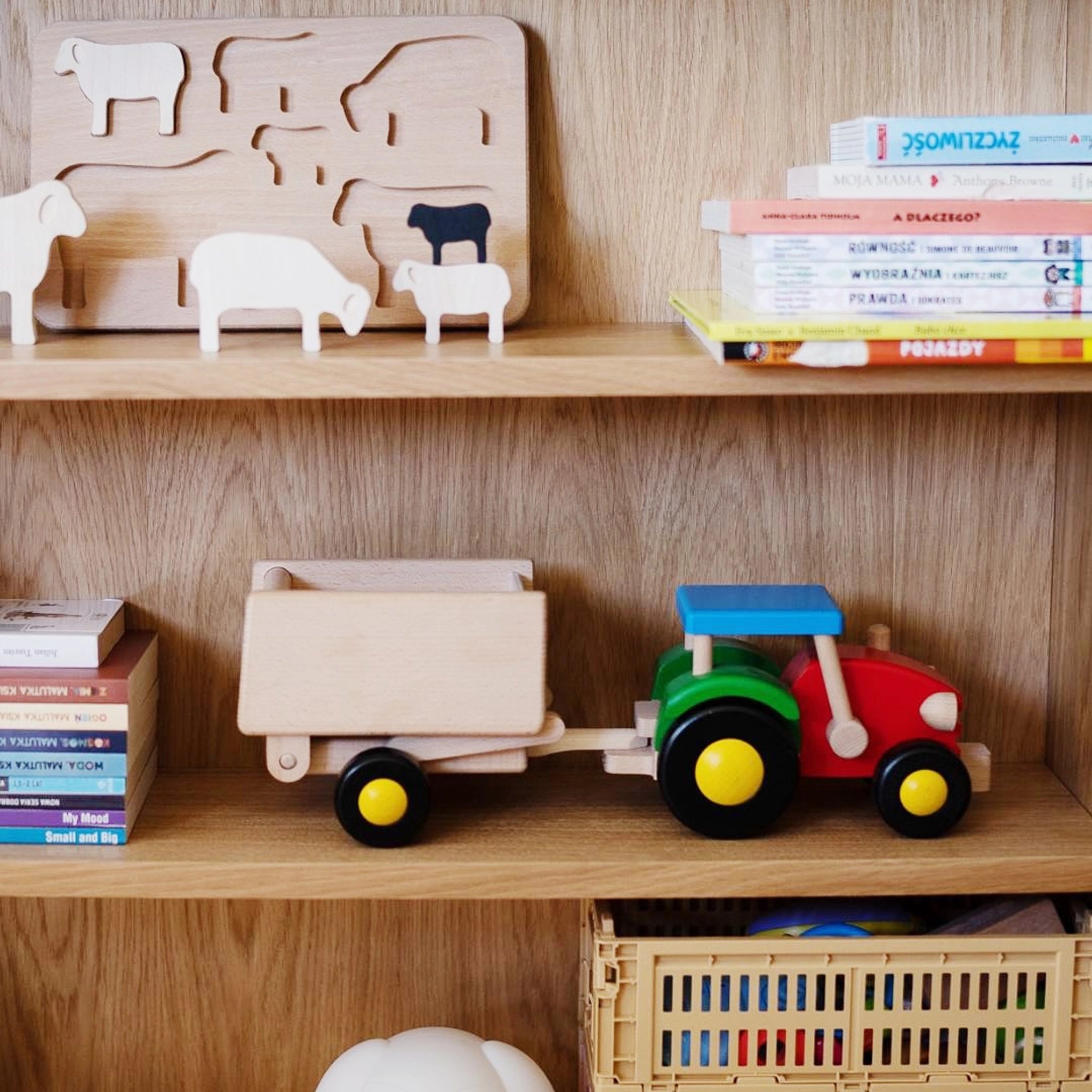 Holzfahrzeug: Traktor mit Anhänger auf Holzregal, umgeben von Holzpuzzle und Kinderbüchern; aus Buchen- und Ahornholz, bunter Traktor mit abnehmbarem Anhänger, handgefertigt in Polen, fördert nachhaltiges Spielen.