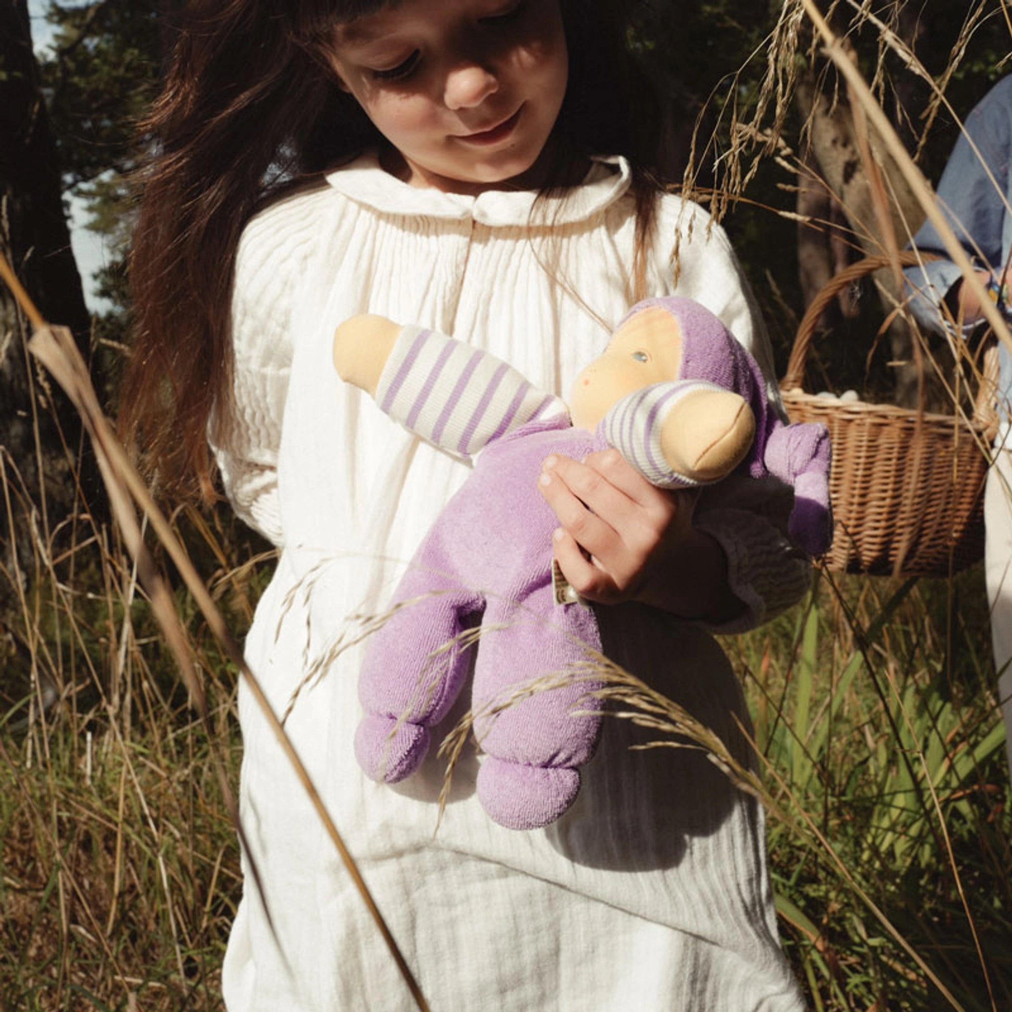 Kind in einem weißen Kleid hält die Puppe: Mein Schatz flieder, eine weiche Puppe aus Bio-Baumwollfrottee mit schadstofffreier Schafschurwolle, ideal zum Kuscheln, in einem rustikalen Feld.