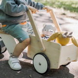 Ein Kind schiebt das Holzfahrzeug: Lastenrad, ein kippsicheres, stabiles Spielzeug aus Buchen- und Ahornholz, mit zwei Kuscheltieren in der Ladefläche, entlang eines Weges.