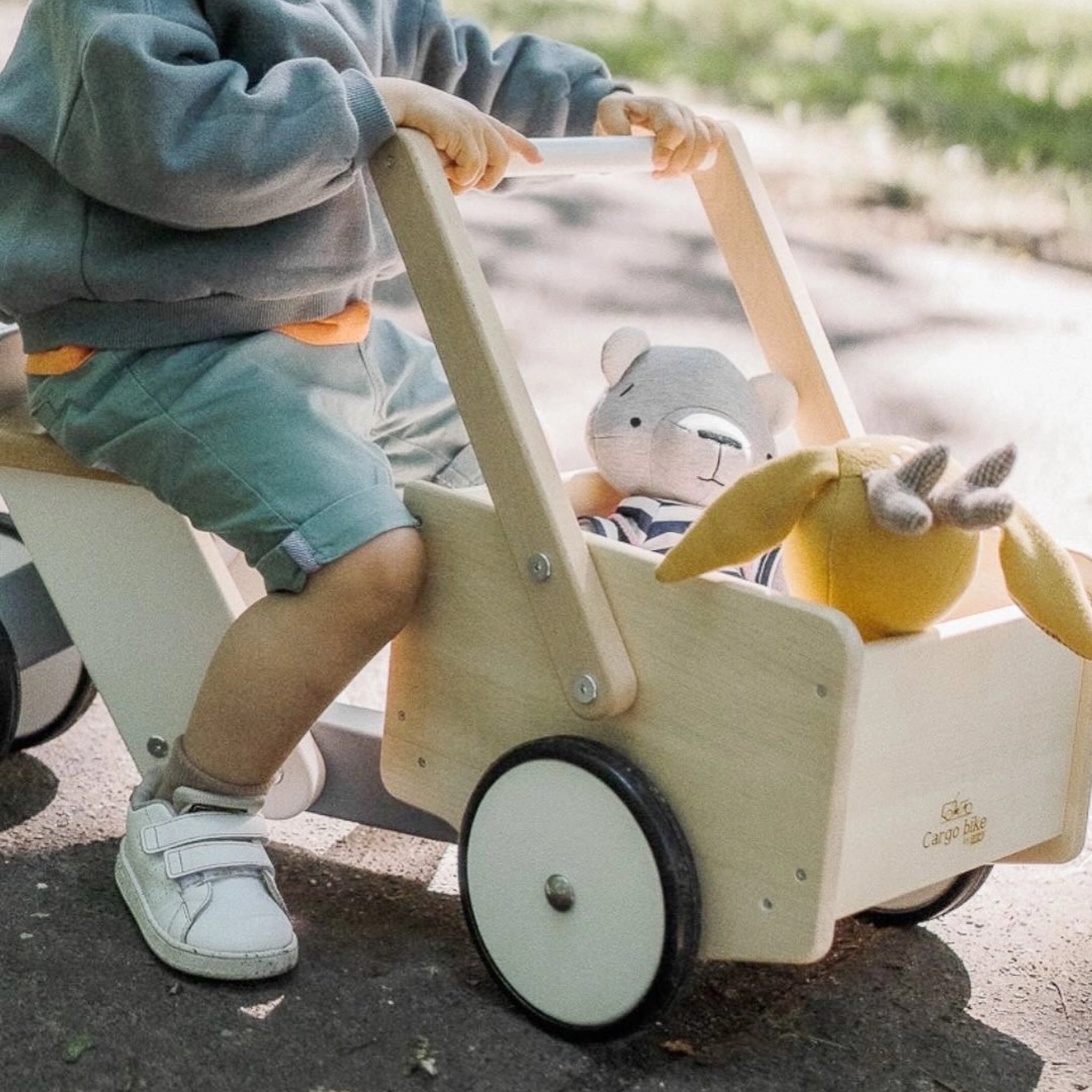Ein Kind schiebt das Holzfahrzeug: Lastenrad, ein kippsicheres, stabiles Spielzeug aus Buchen- und Ahornholz, mit zwei Kuscheltieren in der Ladefläche, entlang eines Weges.
