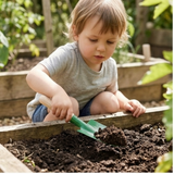 Kleines Kind spielt mit einem Sandschäufelchen aus pulverbeschichtetem Metall in einem Gartenbeet, passend für Kinderhände, ideal für Gartenarbeit. Hergestellt in Deutschland, Teil von Redeckers Sandspielzeug-Kollektion.