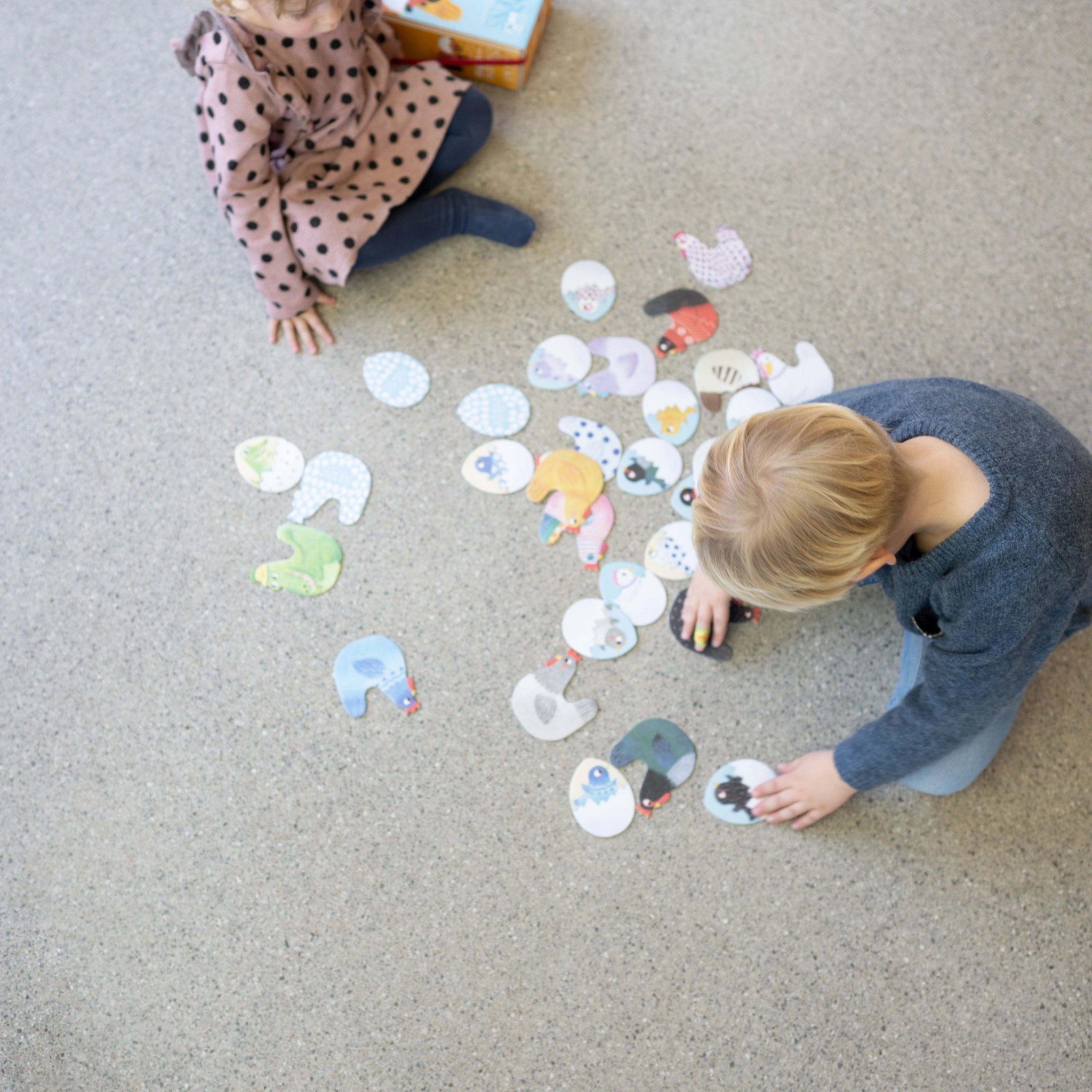 Memory: Chicks & Chickens - Zwei Kinder spielen auf dem Boden mit bunten Papierstücken. Das Spiel fördert Gedächtnis und Konzentration und besteht aus FSC-Papier und recyceltem Karton.