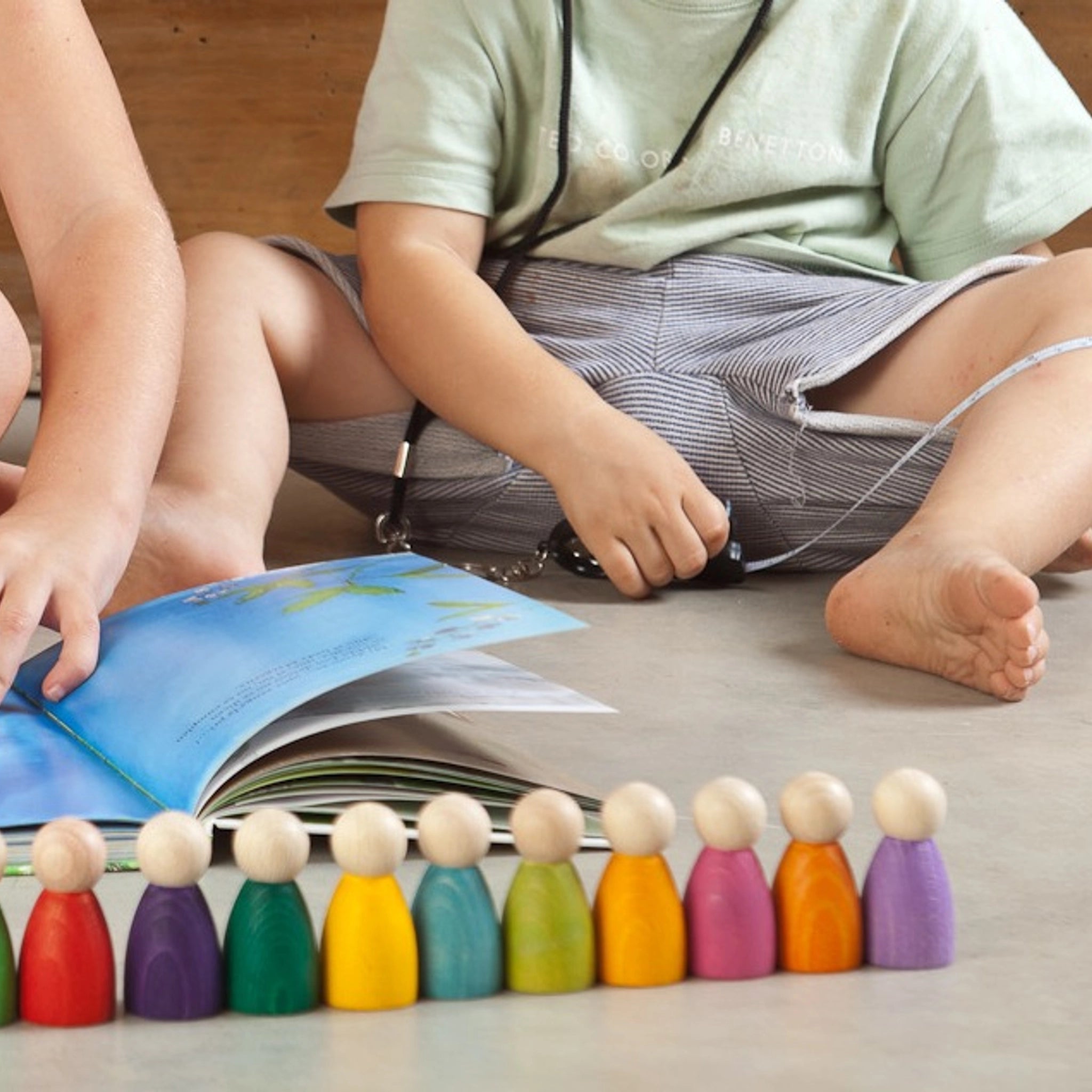 Freies Spiel: 12 Nins, handgefertigte Holzfiguren aus nachhaltiger Forstwirtschaft, ideal für kreatives Kinderspiel. Im Hintergrund sind Kinder zu sehen, die aufmerksam ein Buch lesen.