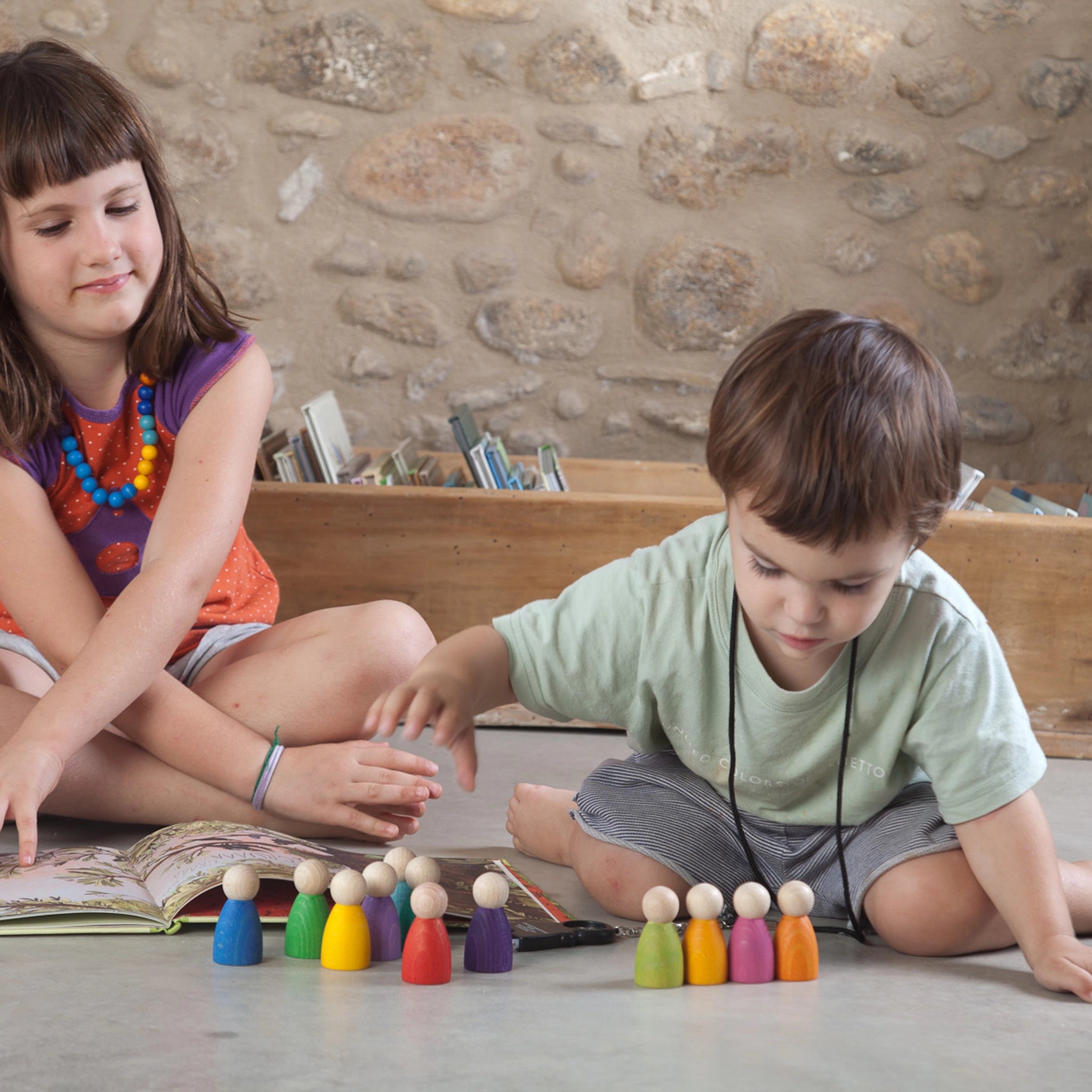 Kinder spielen mit Freies Spiel: 12 Nins, handgefertigten Figuren aus nachhaltig geforstetem Holz, ideal für kreatives Spiel. Ein Junge und ein Mädchen erkunden bunte, regenbogenfarbene Holzfiguren auf einem Bett.